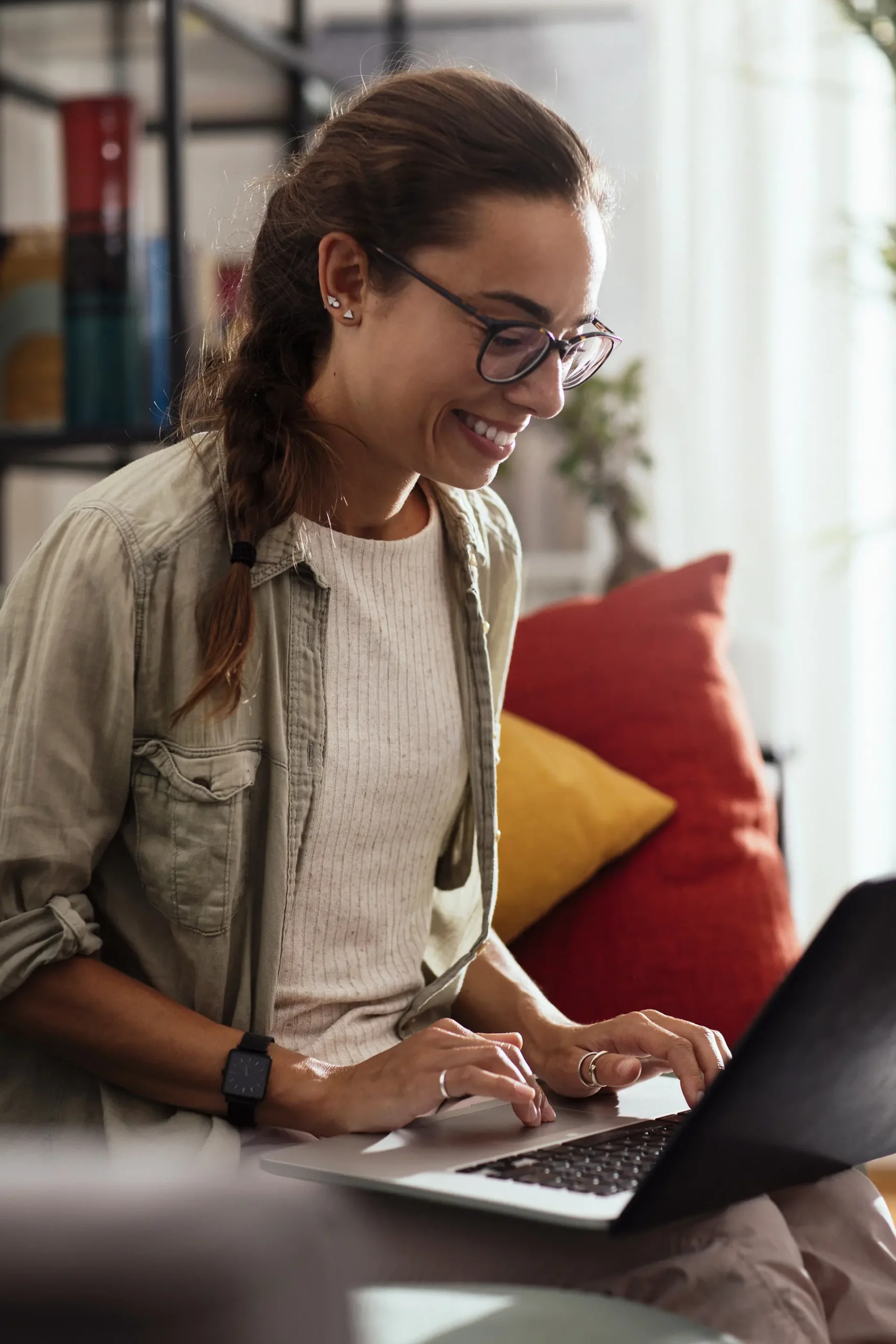 A woman sitting on a couch with red and yellow pillows smiling and working on a laptop.