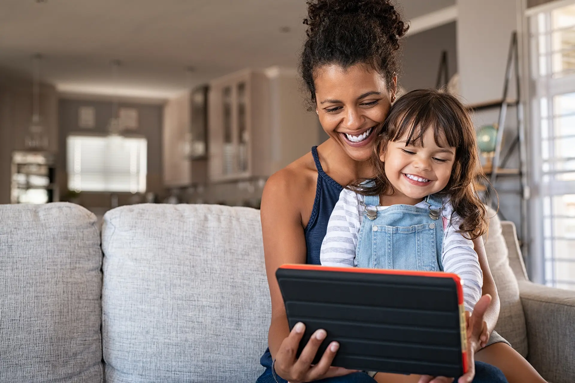 A woman sitting on a gray couch with a young girl in her lap smiling and playing on a tablet computer.