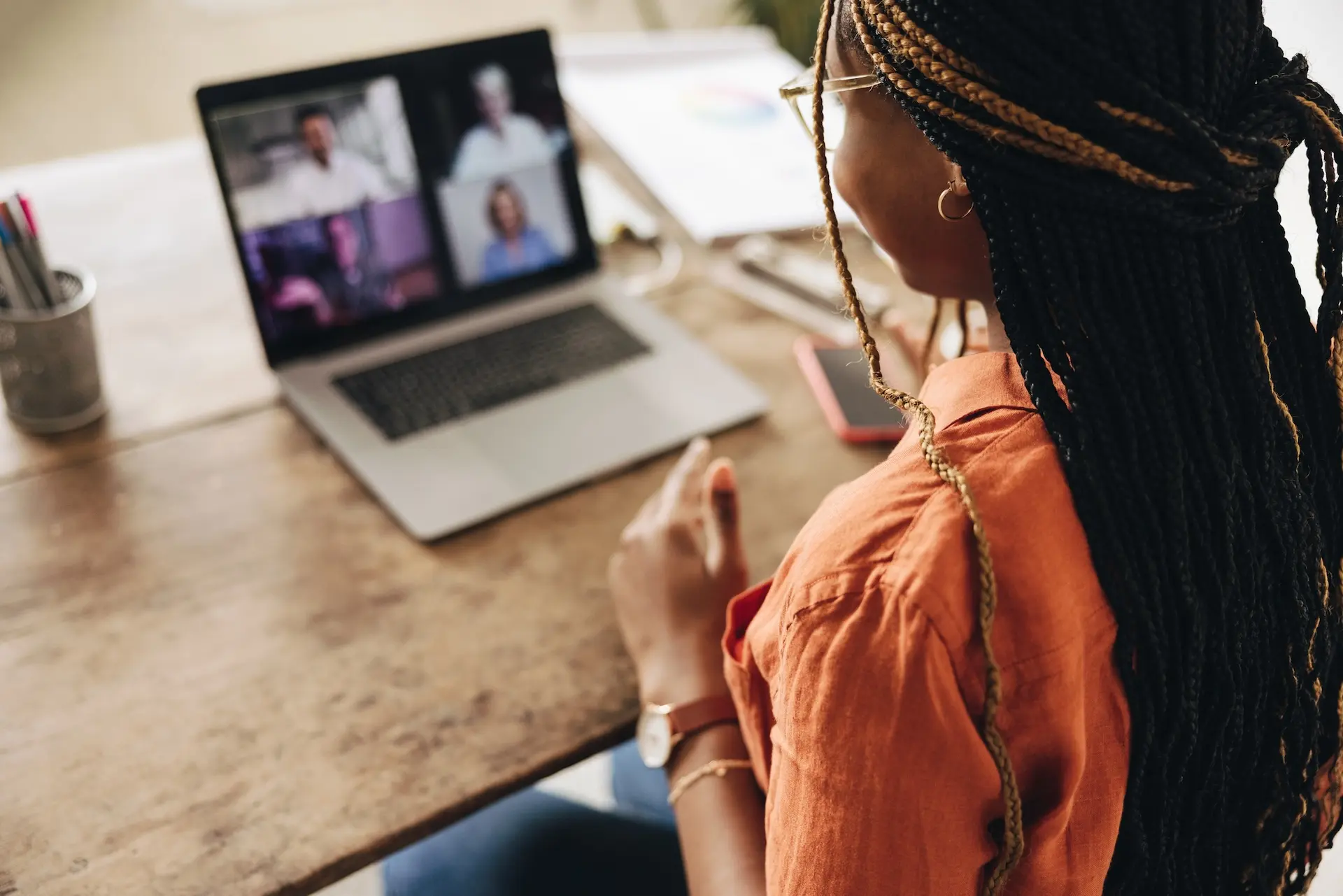 Black woman in an orange button-up shirt participating in a video call with four people on her computer screen.