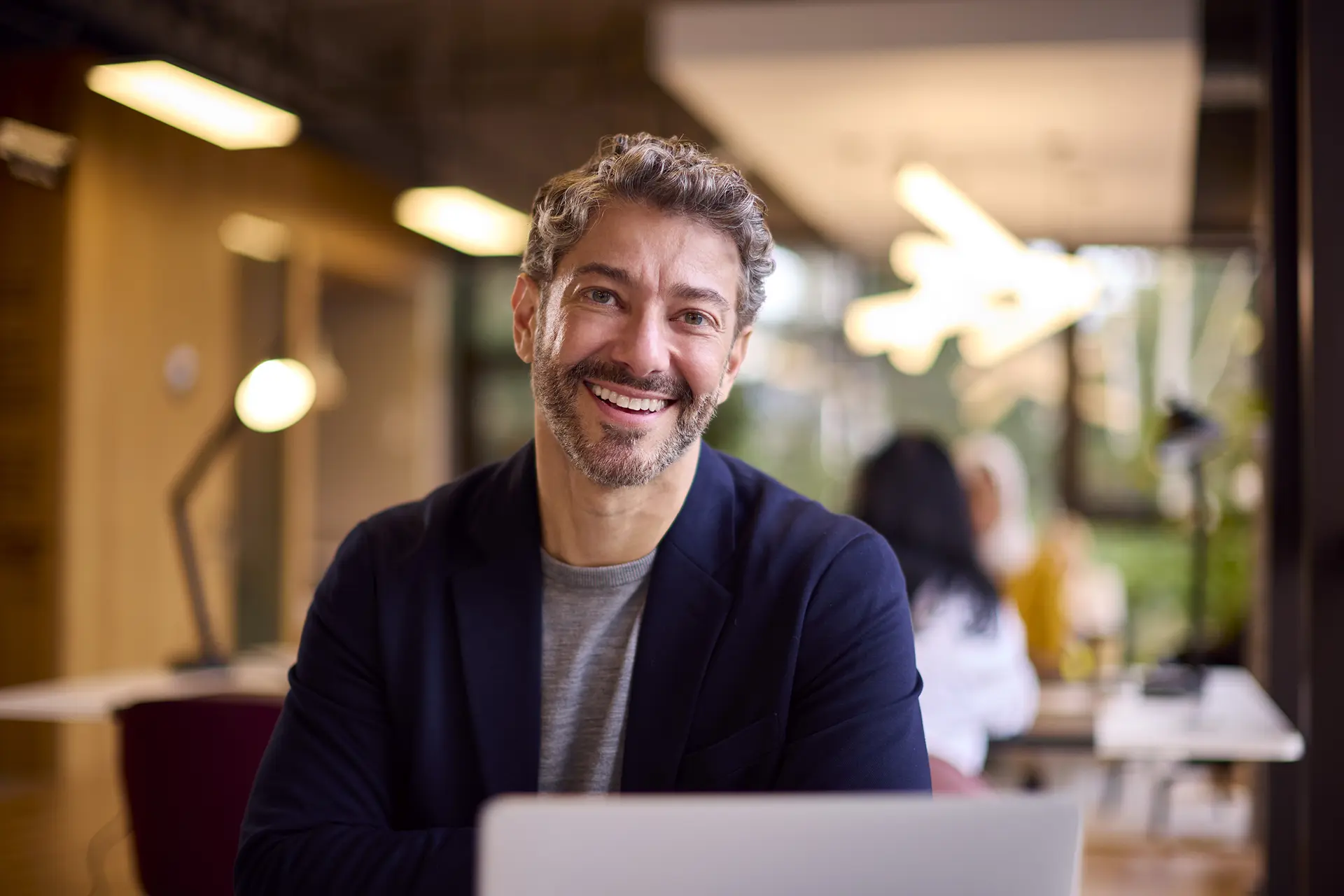 A man in a blue cardigan smiling sitting in front of a computer in a shared workspace with two people working behind him.