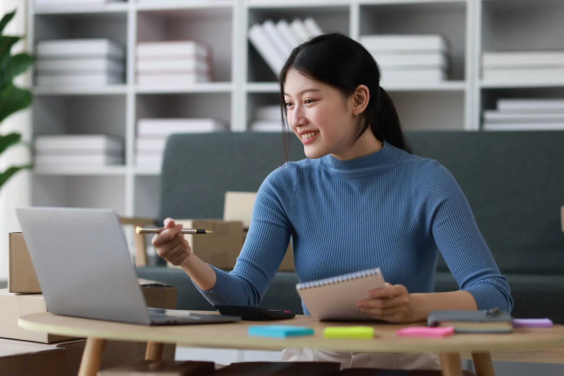 Asian woman in a blue shirt sitting at a desk with a computer taking notes on a spiral pad surrounded by multi-color sticky notes.